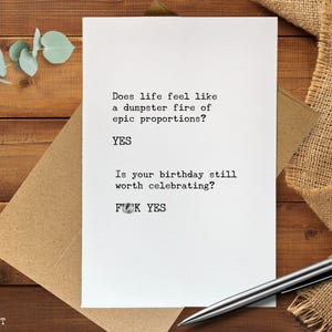 May include: A white greeting card with the text "Does life feel like a dumpster fire of epic proportions? YES. Is your birthday still worth celebrating? FUCK YES." A silver pen rests on the card, which is on a brown envelope.