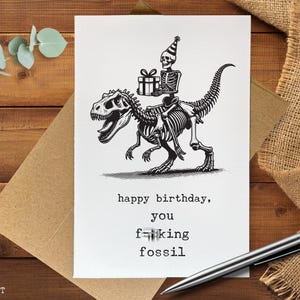 May include: A white greeting card with a black and white illustration of a skeleton riding a dinosaur skeleton, holding a gift. The card reads "happy birthday, you f**king fossil."
