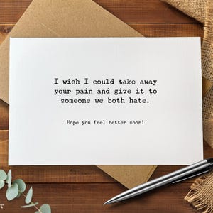 May include: A white greeting card with the text "I wish I could take away your pain and give it to someone we both hate. Hope you feel better soon!" is placed on a wooden surface with a pen and envelope.