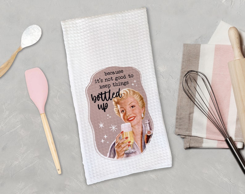 May include: White kitchen towel featuring a vintage-style illustration of a woman holding a glass of bubbly. The text reads "because it's not good to keep things bottled up".