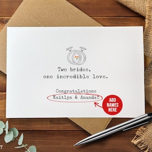May include: A white greeting card with the text "Two brides, one incredible love." and "Congratulations Kaitlyn & Amanda!" is displayed. The card features a drawing of two wedding rings with a rainbow heart. A pen and envelope are also visible.