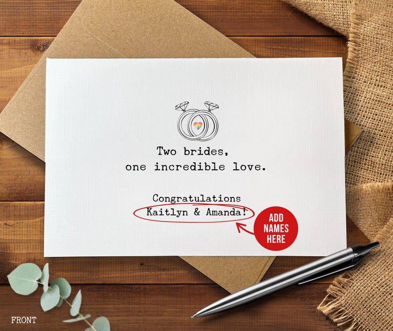 May include: A white greeting card with the text "Two brides, one incredible love." and "Congratulations Kaitlyn & Amanda!" is displayed. The card features a drawing of two wedding rings with a rainbow heart. A pen and envelope are also visible.