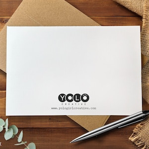 May include: A white card with the text "YOLO GIRL CREATIVE" and a website address, resting on a brown envelope. A silver pen and sprig of greenery are also visible. The card is on a wooden surface.