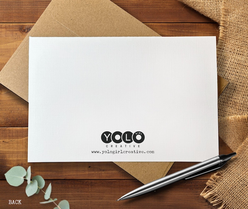 May include: A white card with the text "YOLO GIRL CREATIVE" and a website address, resting on a brown envelope. A silver pen and sprig of greenery are also visible. The card is on a wooden surface.