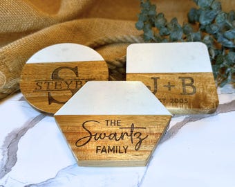 May include: Three wooden coasters with a white marble top. The coasters are engraved with different names and dates. The first coaster says "Steyb" with a large "S". The second coaster says "J+B Est. 2005". The third coaster says "The Schwartz Family".