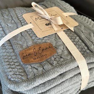 May include: A folded gray cable knit blanket tied with a cream ribbon. A brown leather tag with the names "Liam & Emma" and the date "10.25.2025" is attached. A cream tag with the words "Made with Love" is also attached.