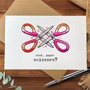 May include: A greeting card with a white background features two pairs of scissors in a crossed design. The scissors have pink, orange, and yellow handles. The text "rock, paper, scissors?" is printed below the scissors. The card is on a wooden surface.