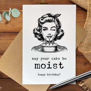 May include: A black and white birthday card featuring a vintage illustration of a woman winking and holding a cake with lit candles. The card reads "may your cake be moist" and "happy birthday!"