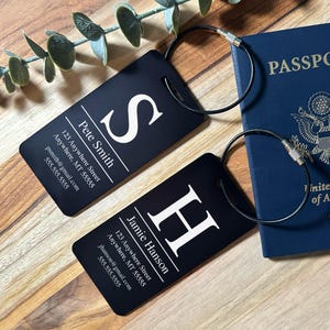 May include: Two black luggage tags with white text and large initial letters, 'S' and 'H'. Each tag has a metal loop and includes a name, address, and contact information. A blue passport is in the background.
