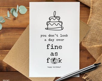 Może przedstawiać: Biała kartka urodzinowa z czarnym rysunkiem tortu i napisem "you don't look a day over fine as f**k happy birthday!". Karta leży na brązowej kopercie z długopisem.