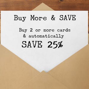 May include: A white card inside a tan envelope with the text "Buy More & SAVE". The card also states "Buy 2 or more cards & automatically SAVE 25%" in a typewriter-style font. The background is a wooden surface.