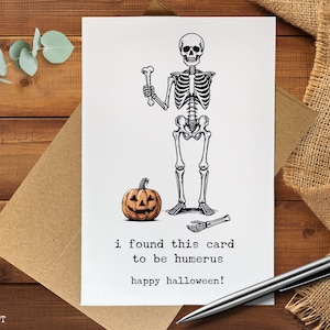 May include: A Halloween greeting card featuring a black and white skeleton illustration holding a bone and standing next to a carved pumpkin. The card reads "i found this card to be humerus happy halloween!"
