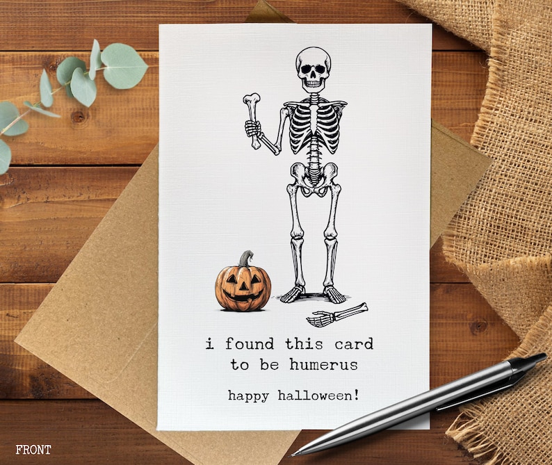 May include: A Halloween greeting card featuring a black and white skeleton illustration holding a bone and standing next to a carved pumpkin. The card reads "i found this card to be humerus happy halloween!"
