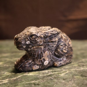 May include: A small, intricately carved rabbit figurine made of dark gray stone with white flecks. The rabbit is sitting with its head tilted to the side.