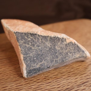 May include: A fragment of pottery with a rough, textured surface. The piece has a light orange-brown color on one side and a dark gray-black color on the other. The pottery shard is resting on a wooden surface.