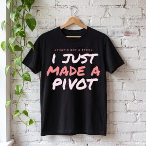 Puede incluir: Una camiseta negra de manga corta con el texto "THAT'S NOT A TYPO" en letras pequeñas de color naranja en la parte superior, seguido de las palabras "I JUST MADE A PIVOT" en letras grandes, negritas, de color rosa claro y coral.