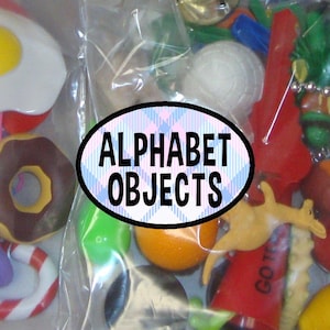 May include: A collection of small plastic miniature objects in a clear bag, including a fried egg, a chocolate donut, a candy cane, an orange, and a kangaroo. An oval label with a blue and white plaid pattern reads ALPHABET OBJECTS.