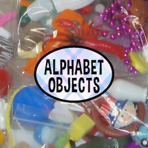 May include: A clear plastic bag filled with various small colorful miniature toys and objects, including a bright orange lobster and purple beads, with an oval label in the center featuring the text ALPHABET OBJECTS in black.