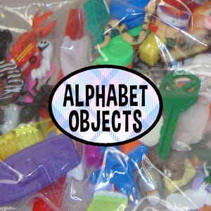 May include: A collection of assorted miniature plastic trinkets and figurines in a clear plastic bag, featuring a green key, a brown donut, a snake, and various colorful shapes. A central oval label displays the text ALPHABET OBJECTS.