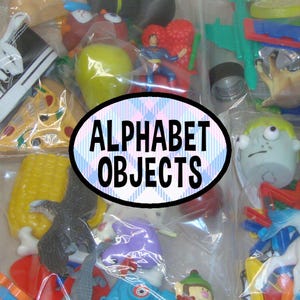 May include: A collection of assorted small plastic toys and figurines packed in clear bags, including a black sneaker, a yellow pear, a Superman figure, a slice of pizza, a corn cob, and a zombie head, with the text ALPHABET OBJECTS in the center.
