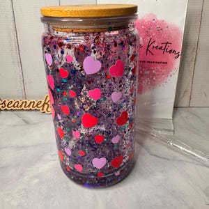 Puede incluir: Un tarro de vidrio transparente con tapa de madera, lleno de purpurina morada y pegatinas en forma de coraz&oacute;n rosas y rojas. El tarro est&aacute; decorado con una variedad de formas de coraz&oacute;n y purpurina, creando un dise&ntilde;o festivo y llamativo.