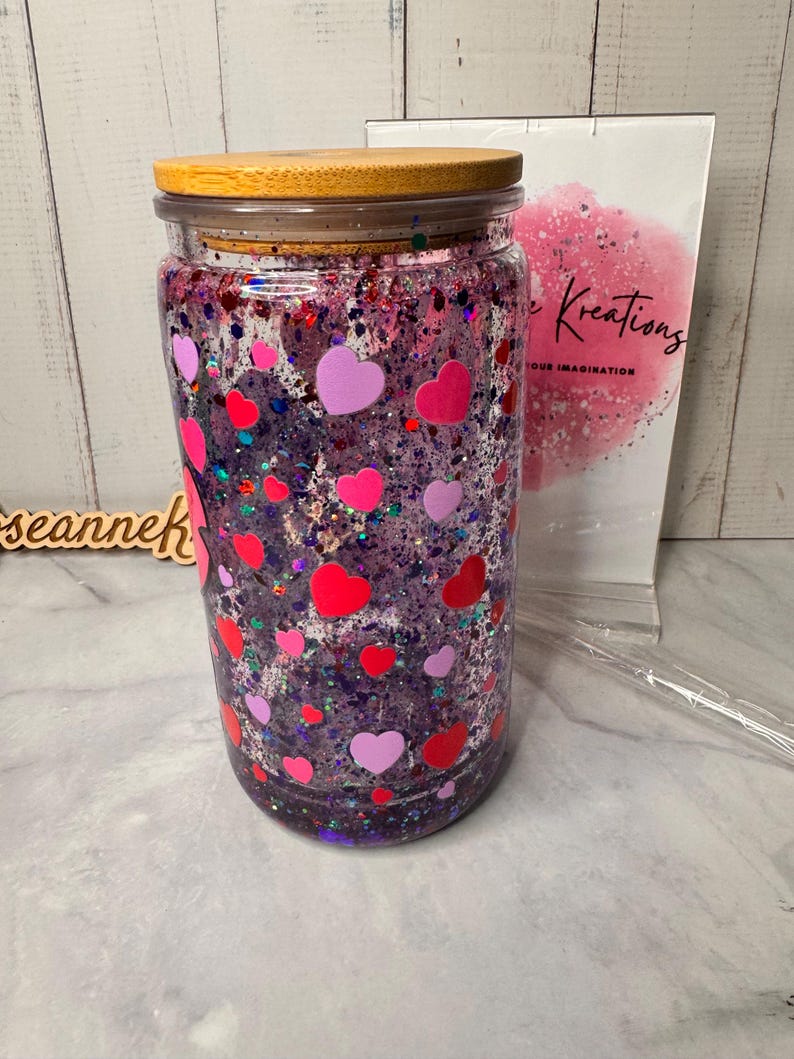 Puede incluir: Un tarro de vidrio transparente con tapa de madera, lleno de purpurina morada y pegatinas en forma de coraz&oacute;n rosas y rojas. El tarro est&aacute; decorado con una variedad de formas de coraz&oacute;n y purpurina, creando un dise&ntilde;o festivo y llamativo.