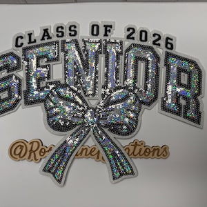 May include: A decorative sign for the graduating class of 2026. The word "SENIOR" is spelled out in large, shimmering letters, with the text "CLASS OF 2026" above. A sequined bow is below the word "SENIOR". The sign is white with black trim.