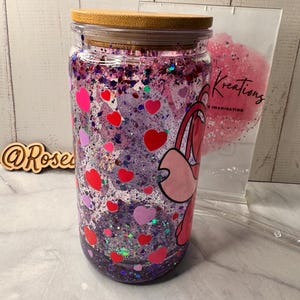 Puede incluir: Un vaso de vidrio transparente con tapa de bamb&uacute;, lleno de purpurina morada, formas de coraz&oacute;n rojas y rosas y un personaje de dibujos animados. El vaso tiene un dise&ntilde;o de coraz&oacute;n rosa y rojo y el texto "Kreations" es visible.