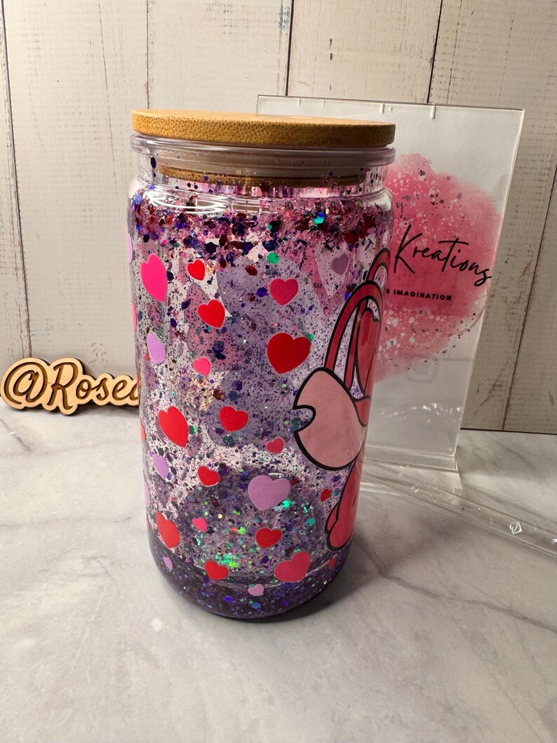 Puede incluir: Un vaso de vidrio transparente con tapa de bamb&uacute;, lleno de purpurina morada, formas de coraz&oacute;n rojas y rosas y un personaje de dibujos animados. El vaso tiene un dise&ntilde;o de coraz&oacute;n rosa y rojo y el texto "Kreations" es visible.