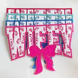 May include: A white felt patch with the word "WIFEY" embroidered in pink. The patch has a pink bow with sequins attached to the bottom.