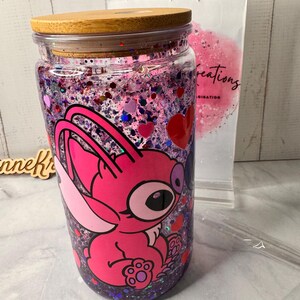 Puede incluir: Un vaso de vidrio transparente con tapa de bamb&uacute;, lleno de purpurina rosa y morada y un personaje de dibujos animados rosa. El vaso tiene una pajita y un soporte de pl&aacute;stico transparente en el fondo.