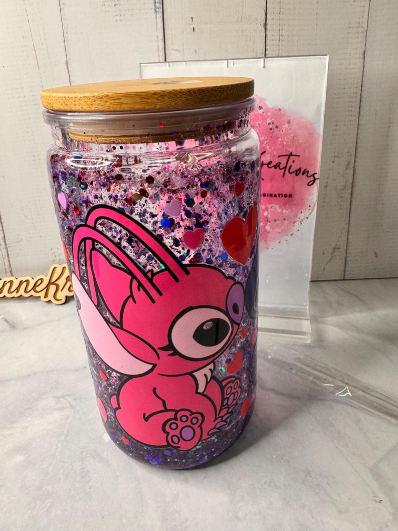 Puede incluir: Un vaso de vidrio transparente con tapa de bamb&uacute;, lleno de purpurina rosa y morada y un personaje de dibujos animados rosa. El vaso tiene una pajita y un soporte de pl&aacute;stico transparente en el fondo.