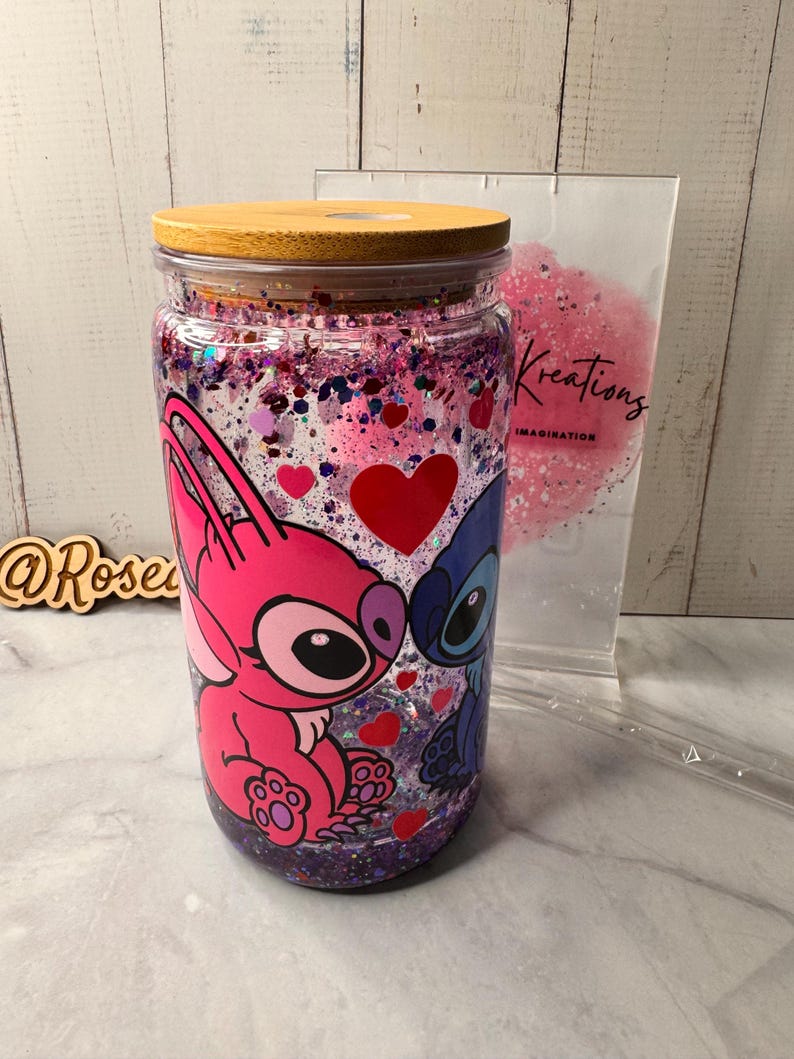 Puede incluir: Un vaso de vidrio transparente con tapa de madera, con un dise&ntilde;o de personajes de dibujos animados rosas y azules con corazones. El vaso est&aacute; lleno de purpurina y confeti peque&ntilde;o en forma de coraz&oacute;n. Una bolsa transparente con la palabra "Kreations" est&aacute; en el fondo.