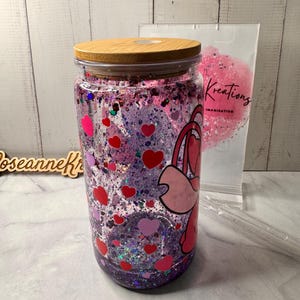 Puede incluir: Un tarro de cristal transparente con tapa de madera, lleno de purpurina rosa y morada, corazones rojos y rosas y un personaje de dibujos animados. El tarro tiene una combinaci&oacute;n de colores rosa y morado. El tarro est&aacute; junto a una bolsa que dice "Kreations Imagination".