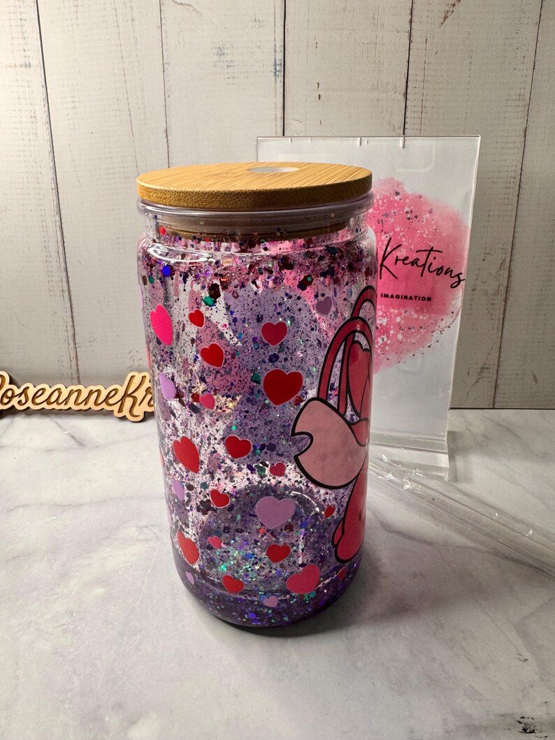 Puede incluir: Un tarro de cristal transparente con tapa de madera, lleno de purpurina rosa y morada, corazones rojos y rosas y un personaje de dibujos animados. El tarro tiene una combinaci&oacute;n de colores rosa y morado. El tarro est&aacute; junto a una bolsa que dice "Kreations Imagination".