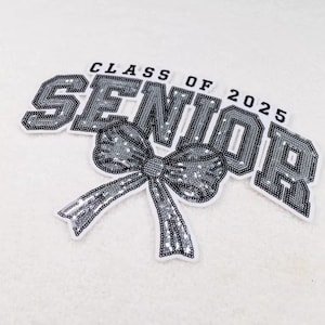 May include: A sequined patch with the words "CLASS OF 2025 SENIOR" in a bold, blocky font. A large bow with flowing ribbons is centered below the word "SENIOR". The patch is primarily silver and white.