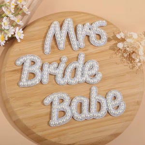 May include: Three white pearl-embellished patches with the words "Mrs", "Bride", and "Babe" on a wooden surface.