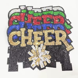 May include: A stack of sequined patches spelling "CHEER" in various colors: gold, blue, red, green, and white. The letter "M" is in black sequins, with a gold and white sequined pom-pom design.
