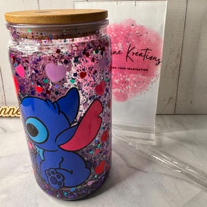 Puede incluir: Un vaso de vidrio transparente con tapa de bamb&uacute;, lleno de purpurina morada y confeti en forma de coraz&oacute;n. Un personaje de dibujos animados azul est&aacute; impreso en la parte delantera. Una pajita de pl&aacute;stico transparente y una bolsa blanca con el texto "ne Kreations" est&aacute;n en el fondo.
