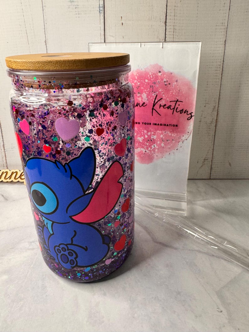 Puede incluir: Un vaso de vidrio transparente con tapa de bamb&uacute;, lleno de purpurina morada y confeti en forma de coraz&oacute;n. Un personaje de dibujos animados azul est&aacute; impreso en la parte delantera. Una pajita de pl&aacute;stico transparente y una bolsa blanca con el texto "ne Kreations" est&aacute;n en el fondo.