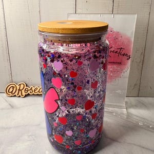 Puede incluir: Un tarro de vidrio transparente con tapa de madera, lleno de purpurina morada, confeti en forma de coraz&oacute;n y un dise&ntilde;o de personaje de dibujos animados. El tarro es de estilo vaso, adecuado para bebidas fr&iacute;as.