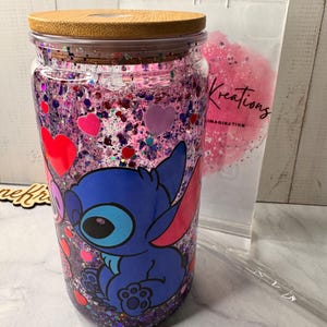 Puede incluir: Un vaso de vidrio transparente con tapa de bamb&uacute;, lleno de purpurina rosa y morada y confeti en forma de coraz&oacute;n. Un personaje de dibujos animados azul aparece en la parte delantera. Tambi&eacute;n se ven una pajita de pl&aacute;stico transparente y una bolsa con la palabra "Kreations".