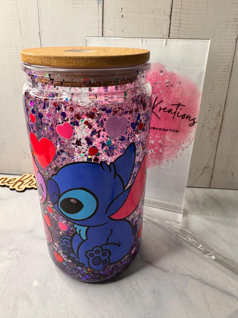 Puede incluir: Un vaso de vidrio transparente con tapa de bamb&uacute;, lleno de purpurina rosa y morada y confeti en forma de coraz&oacute;n. Un personaje de dibujos animados azul aparece en la parte delantera. Tambi&eacute;n se ven una pajita de pl&aacute;stico transparente y una bolsa con la palabra "Kreations".