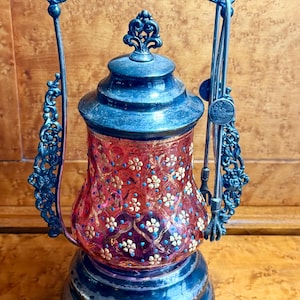Pode incluir: Uma lanterna de vidro antiga com um padrão floral vermelho e dourado. A lanterna tem uma estrutura e alça de metal azul escuro. A base e a tampa também são azul escuro. É uma peça decorativa ornamentada.