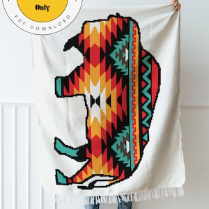 May include: A white crochet blanket with a colorful geometric design of a buffalo. The blanket is held up by a person wearing blue jeans and black boots. The text "Happy Grapher Crochet Pattern Only PDF Download" is on a yellow circle in the top left corner of the image.
