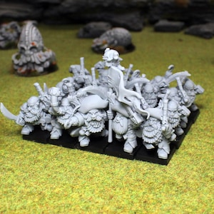 May include: A collection of miniature fantasy figures, likely for tabletop gaming, are positioned on a black base. The figures are detailed with beards, armour, and weapons, set against a backdrop of green terrain and grey rock formations.