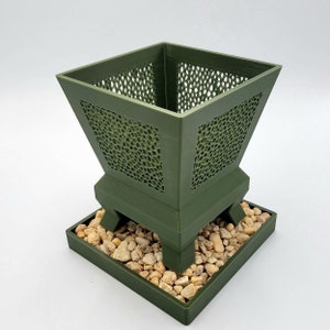 May include: A green ceramic planter with a geometric pattern and a square base. The planter is filled with small, light brown stones.