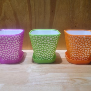 May include: Three plastic flower pots with a textured design. The pots are purple, green, and orange.