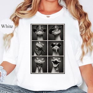 May include: A white t-shirt featuring a black and white graphic print of six snakes in a grid. Each snake is depicted with different accessories, including cowboy hats, a bandana, sunglasses, a bolo tie, and a sheriff star badge.