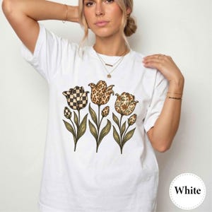Może przedstawiać: Biały t-shirt z krótkim rękawem z nadrukiem trzech tulipanów z zielonymi łodygami i liśćmi. Główki tulipanów mają różne wzory: jeden w czarno-białą szachownicę, jeden w brązowe cętki lamparta, a jeden w drobny kwiatowy wzór.